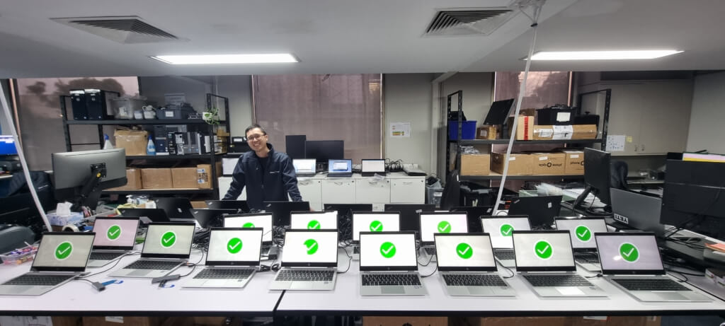 Technician standing behind a row of laptops showing successful data erasure, part of PhoneCycle’s responsible IT asset disposal process.